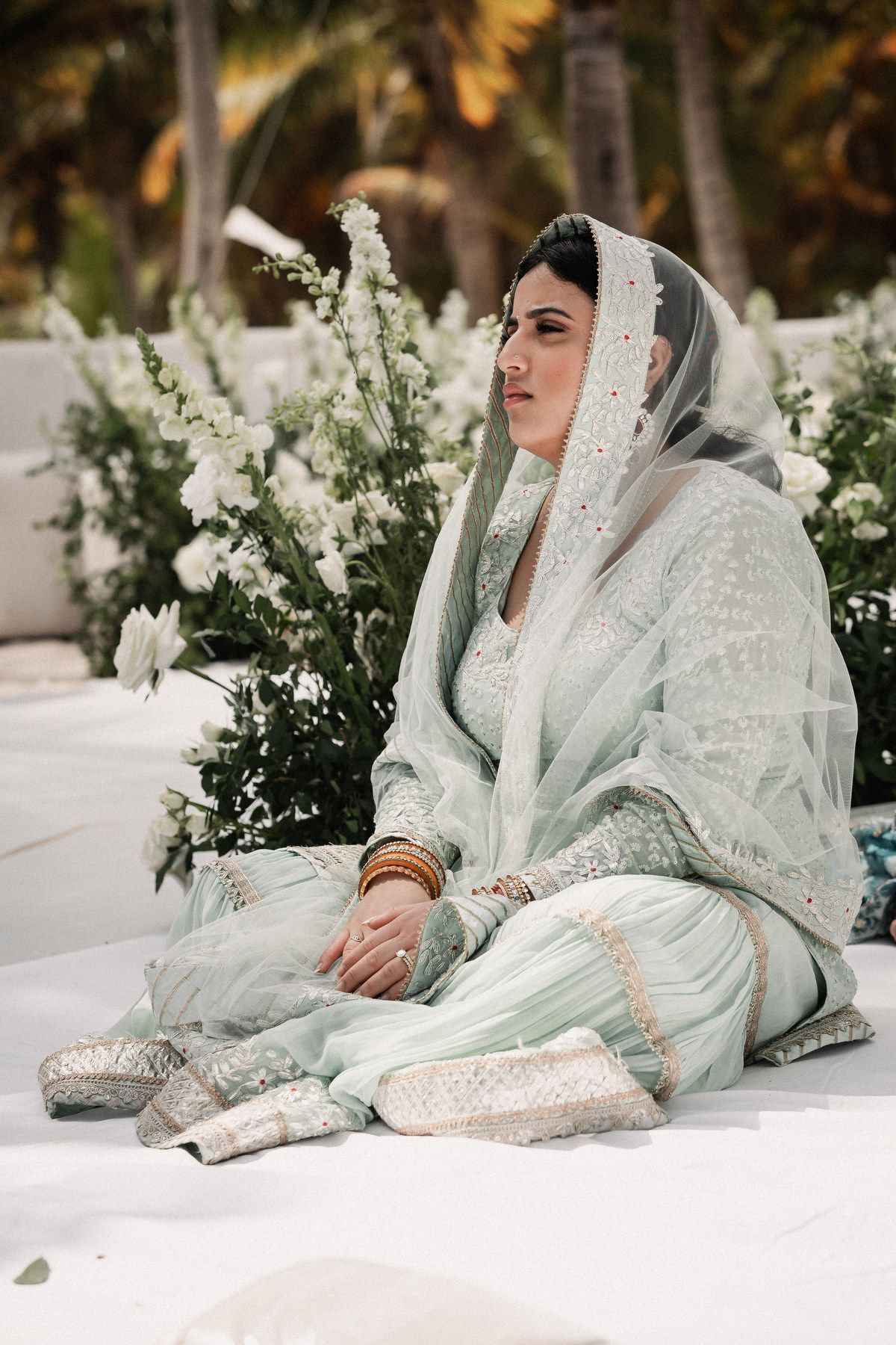 Indian bride in mint green embroidered lehenga with dupatta seated during outdoor beach wedding ceremony.