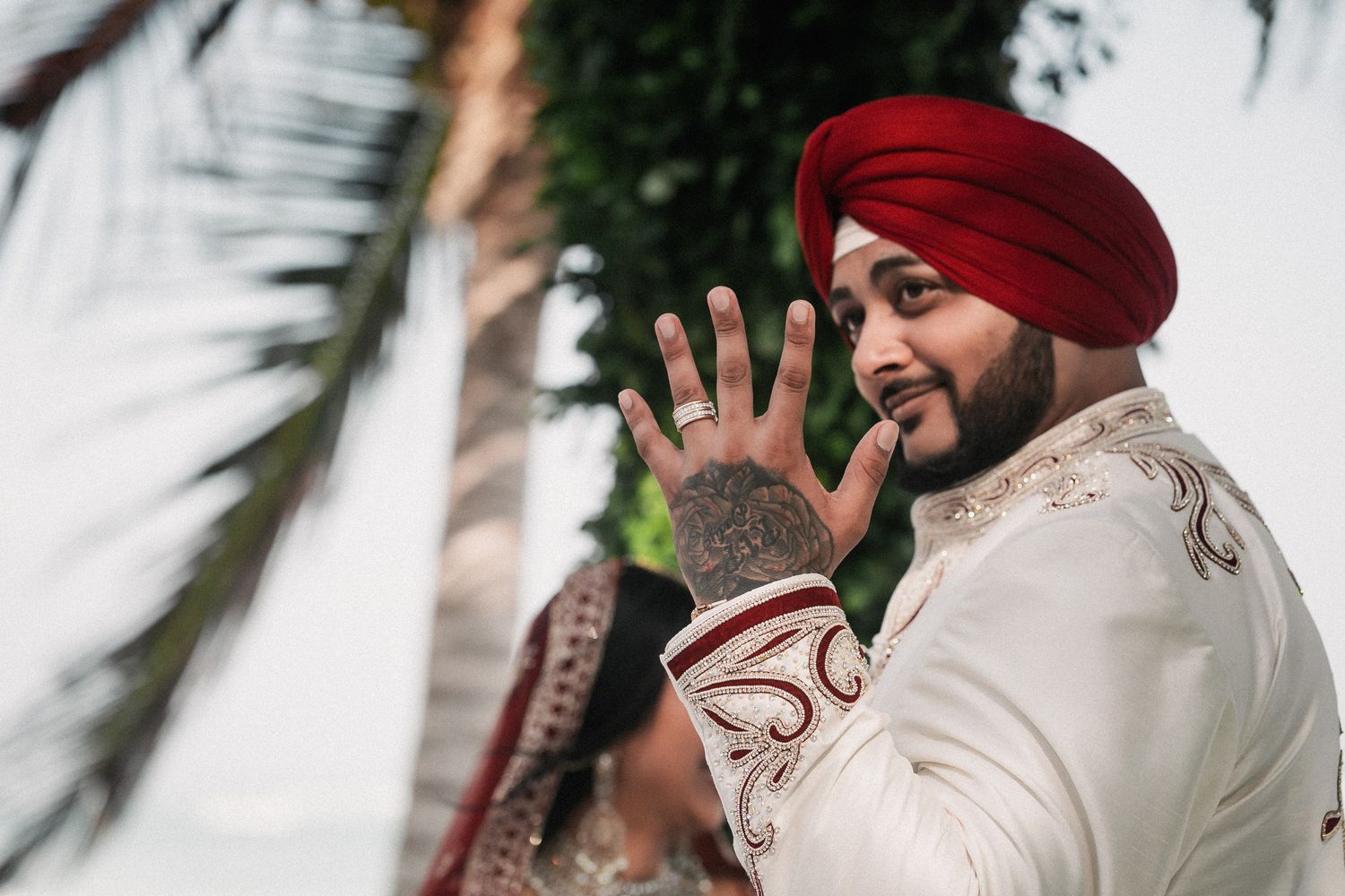 Sikh groom in red turban and white sherwani showing wedding ring to camera at outdoor ceremony.