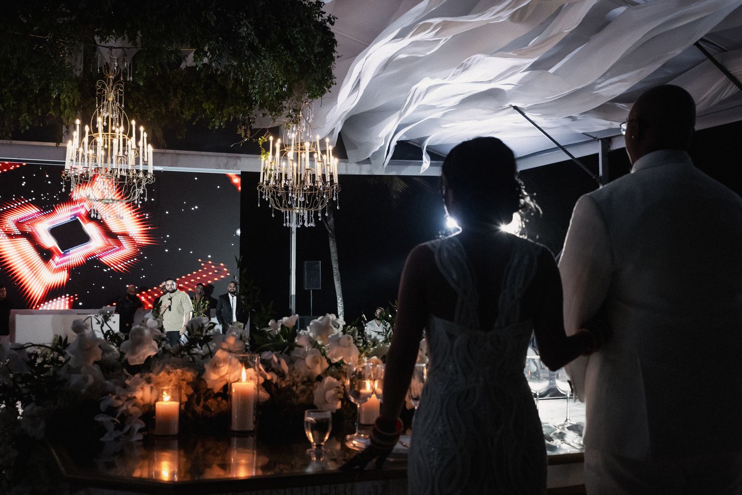Bride and groom silhouetted against glowing candles and chandelier at elegant evening wedding reception.