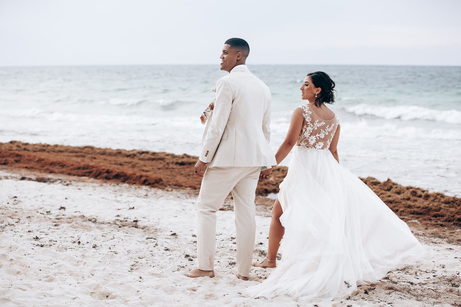 Newlyweds walking along the sandy beach with ocean waves in the background.