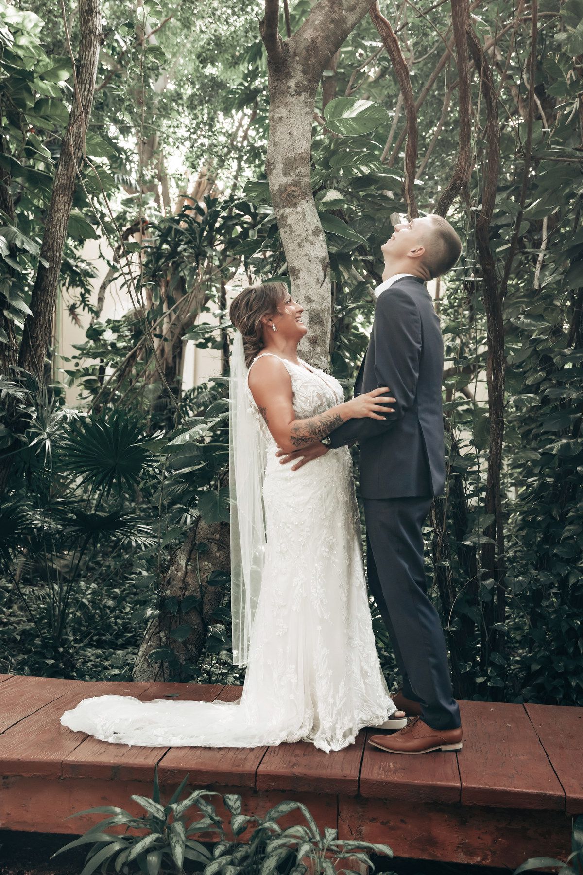 Bride and groom share romantic moment on wooden deck surrounded by lush tropical greenery during outdoor wedding ceremony.
