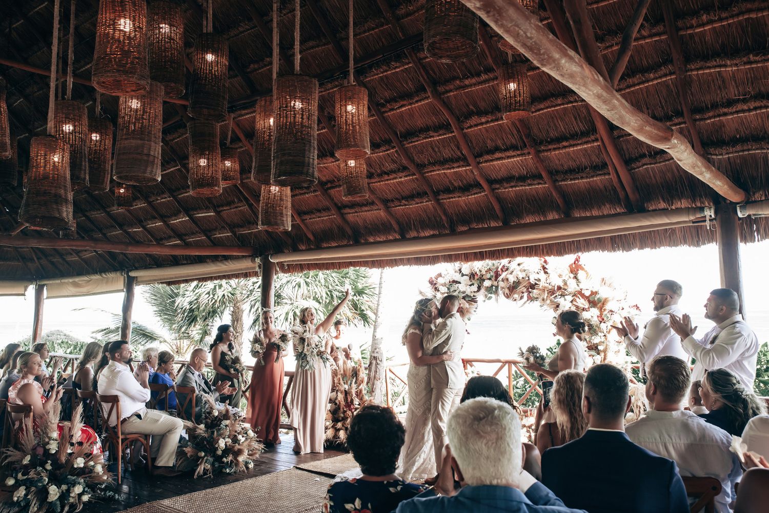 Romantic tropical wedding ceremony under rustic thatched roof with hanging lanterns and floral arch.