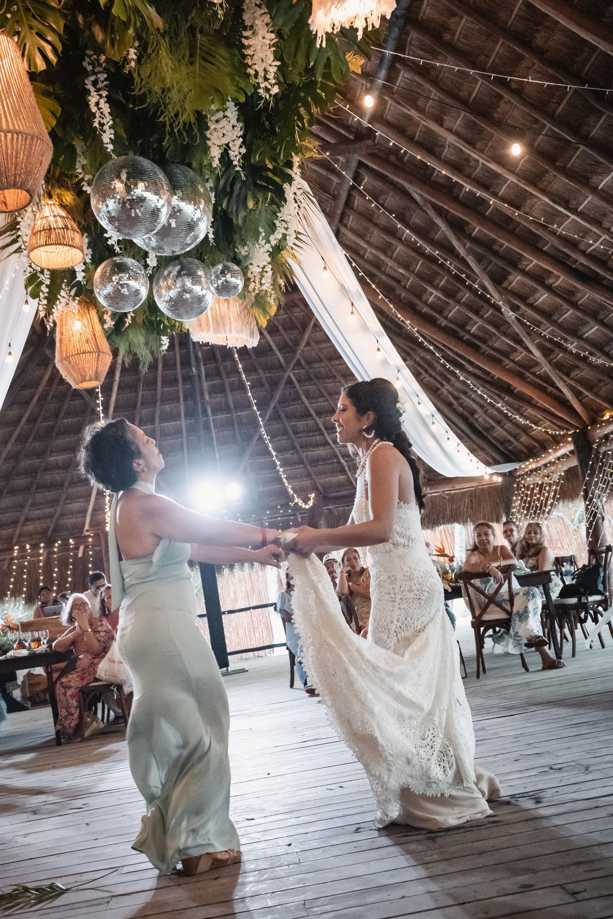 Two brides share a joyful first dance under disco balls and tropical greenery in a thatched-roof wedding venue.