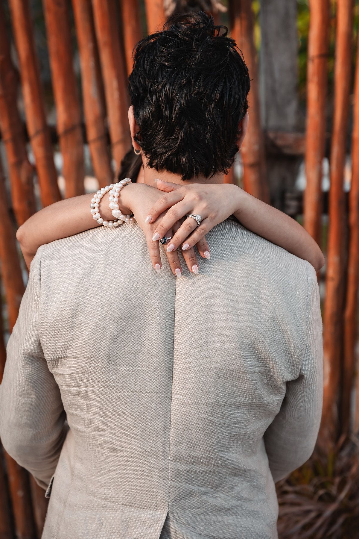 Bride with pearl bracelet and ring embraces groom from behind near rustic bamboo fence.
