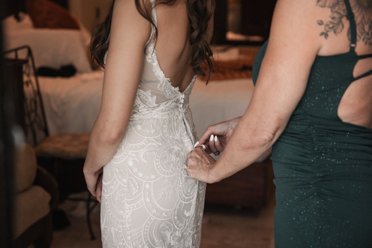 Bridesmaid fastening buttons on brides lace backless wedding gown before ceremony.