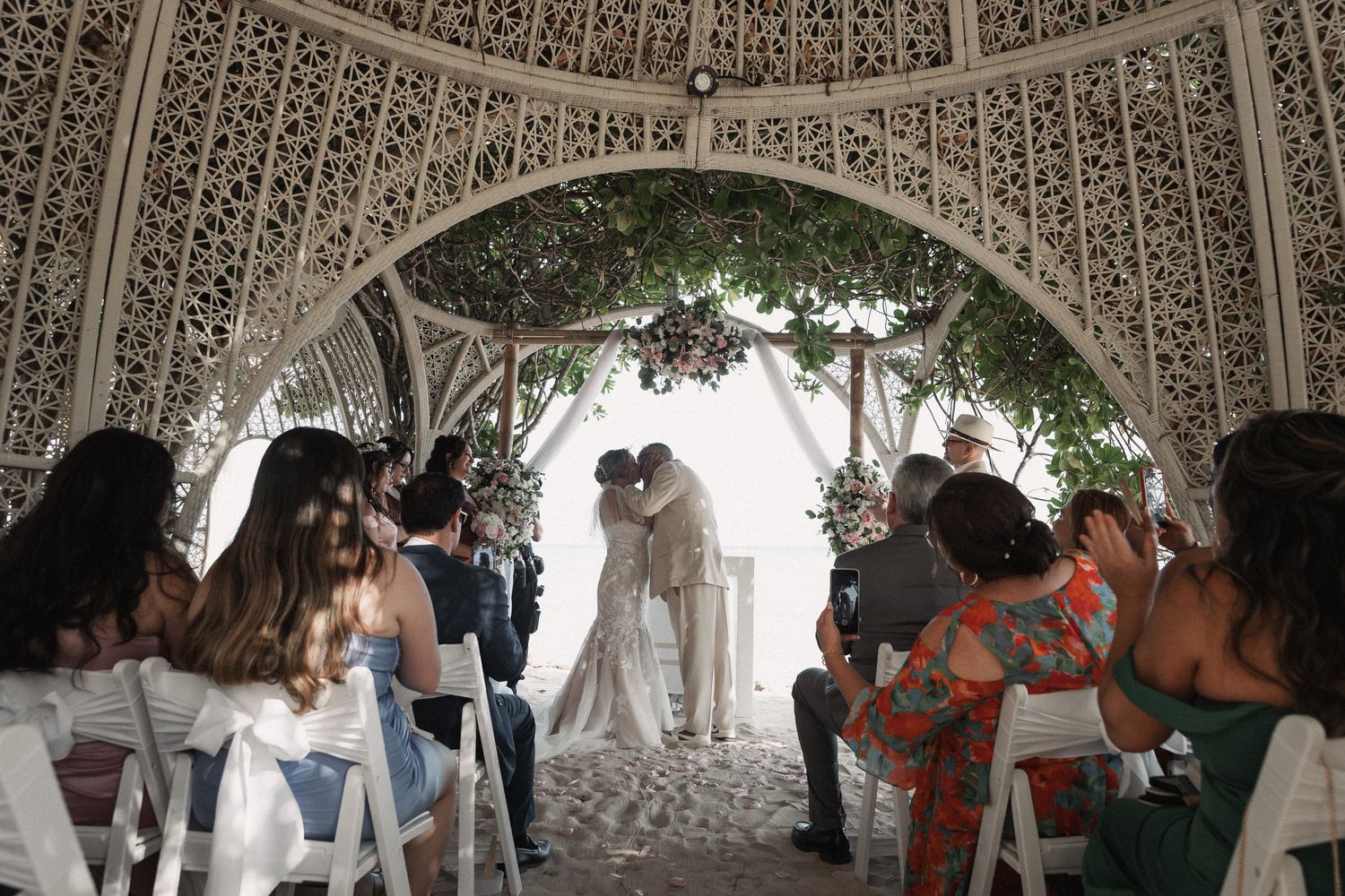 Bride and groom sharing first kiss under ornate lattice arch surrounded by wedding guests.