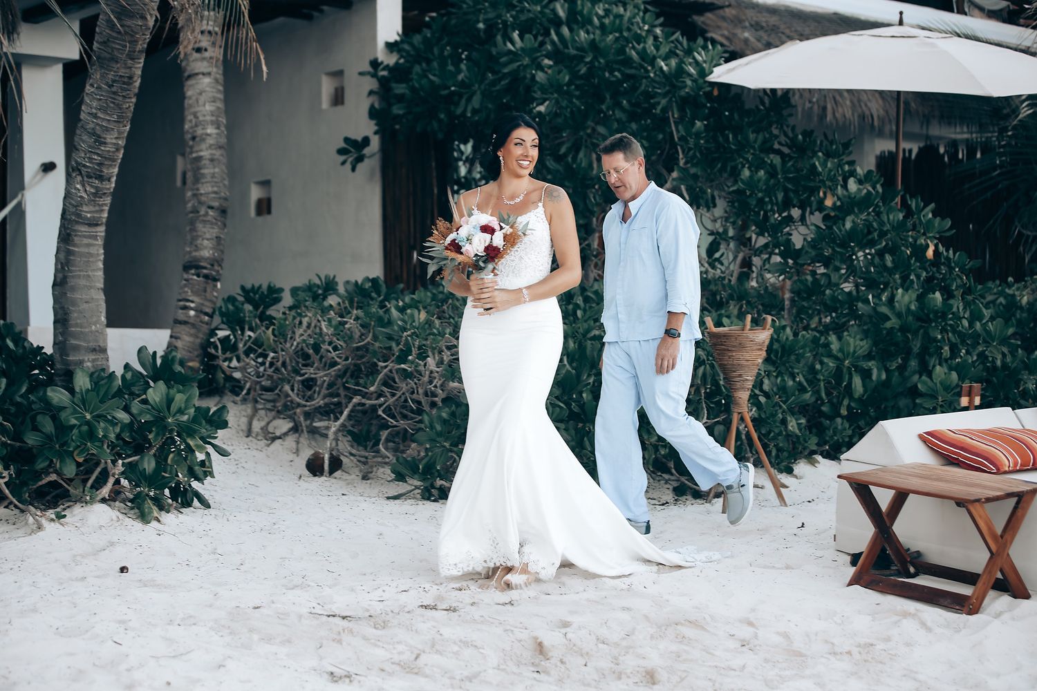 A bride and groom walk together on a beach surrounded by tropical foliage.