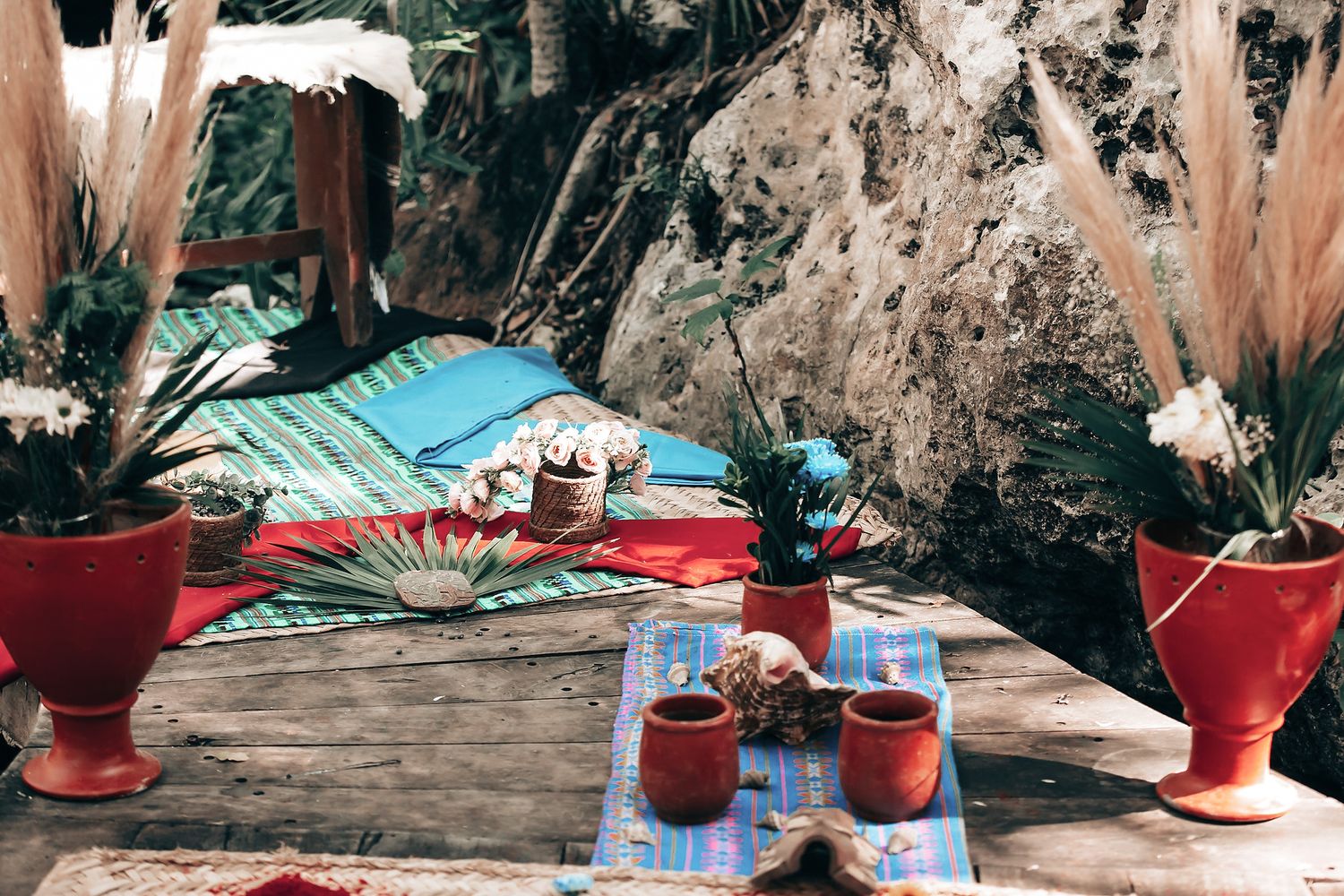 A cozy outdoor setup with a colorful blanket, pillows, and drinks on a wooden table surrounded by potted plants.