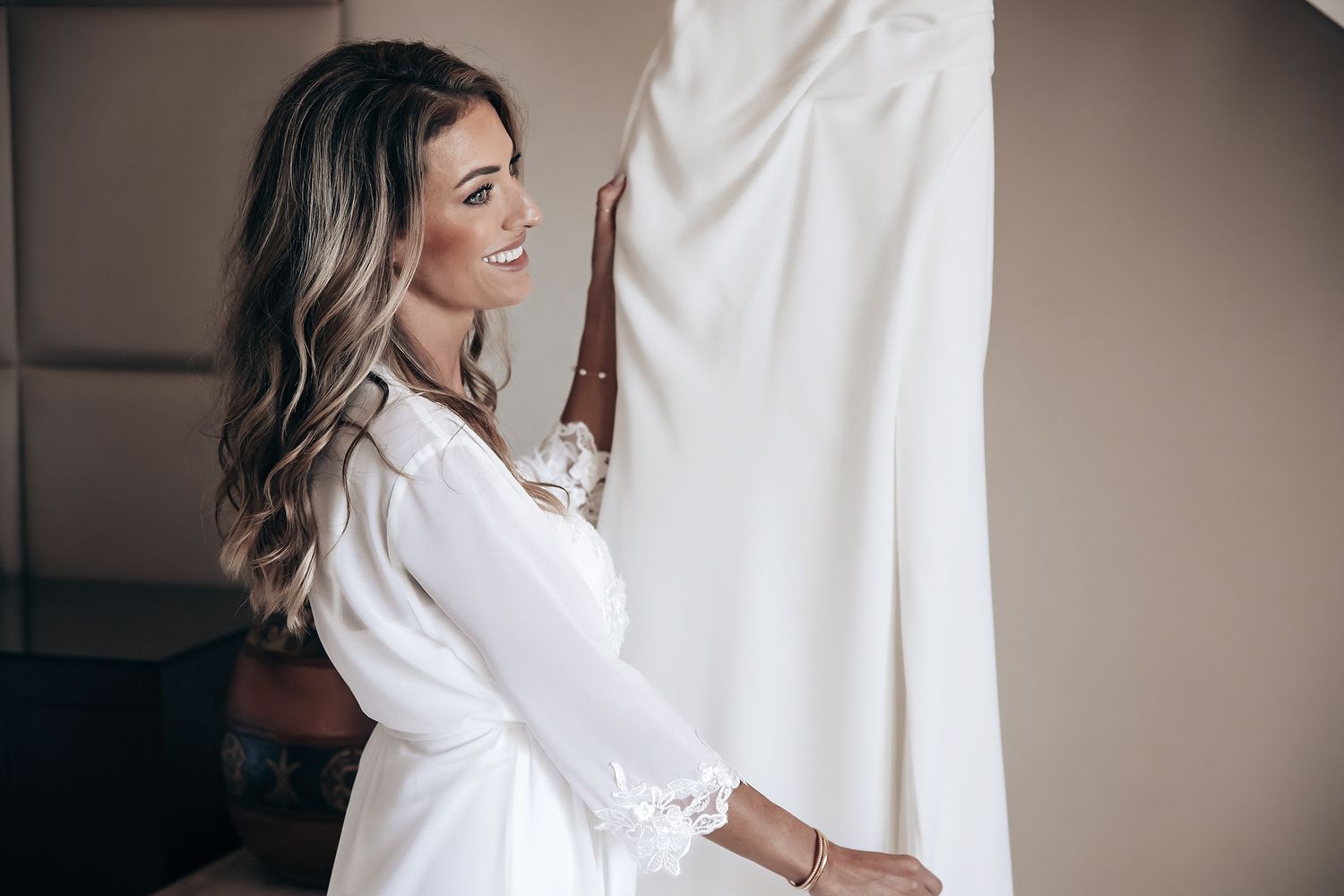 A woman in a white robe stands next to a window, looking at a white wedding dress.