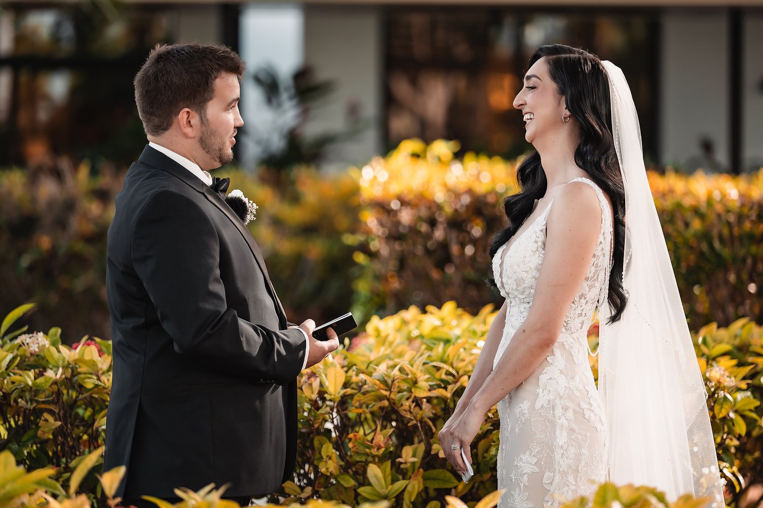 Smiling bride in lace gown and veil listens to groom reading vows at outdoor tropical wedding.