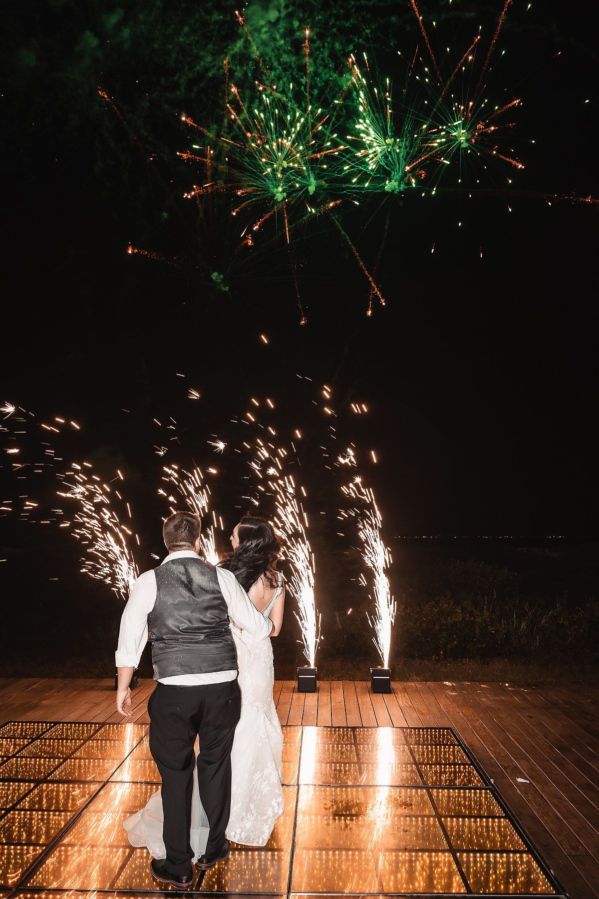 Bride and groom standing on dance floor watching colorful wedding fireworks display lighting up the night sky.