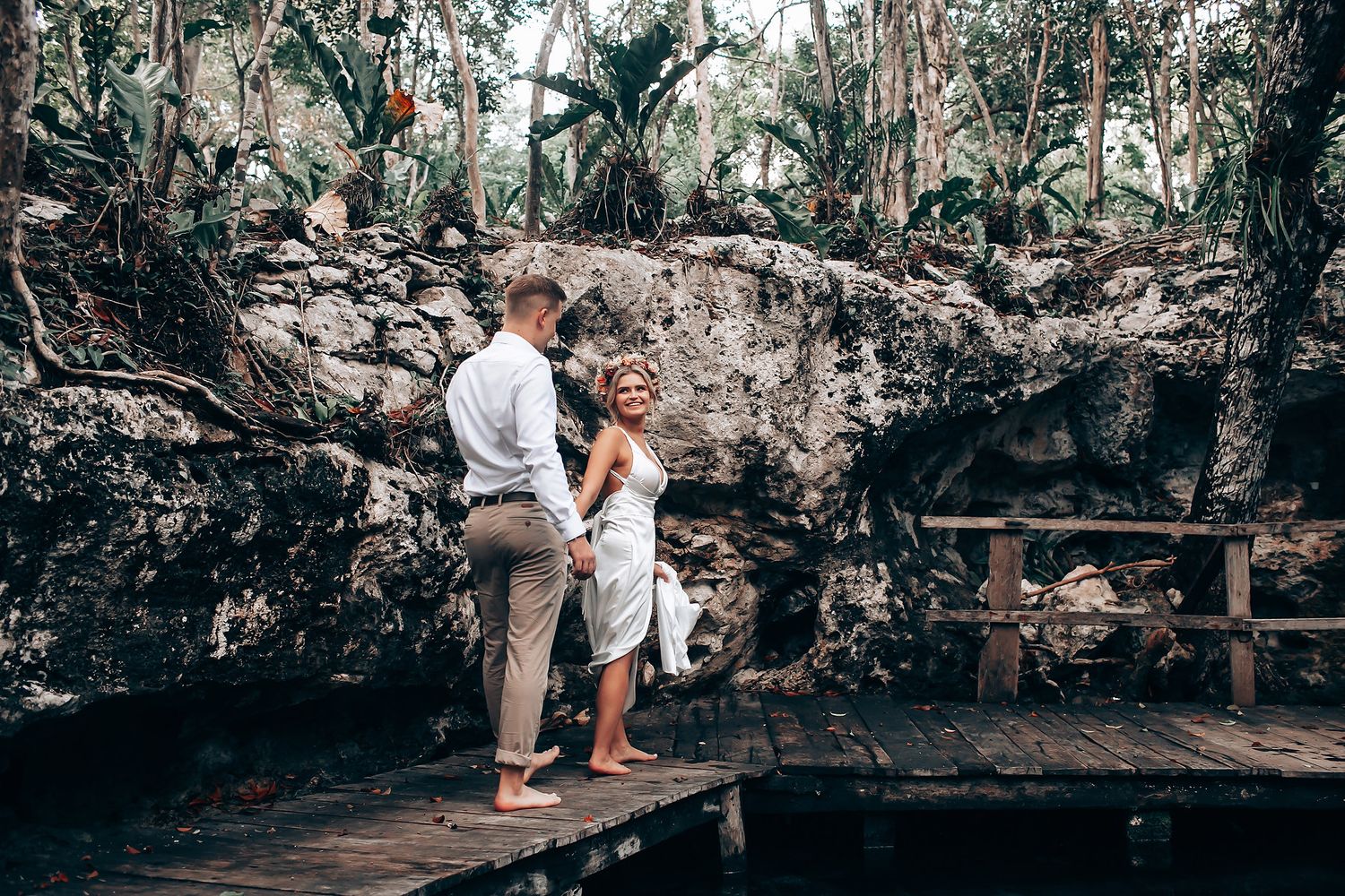 A couple stands together on a wooden deck surrounded by tropical foliage and rocky formations.