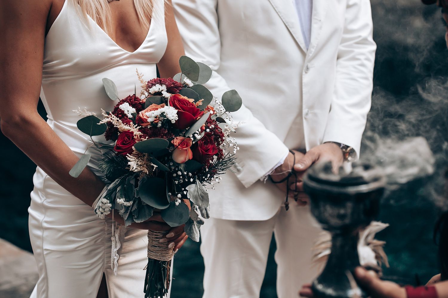 Wedding party members holding deep red and burgundy floral bouquets against white attire.