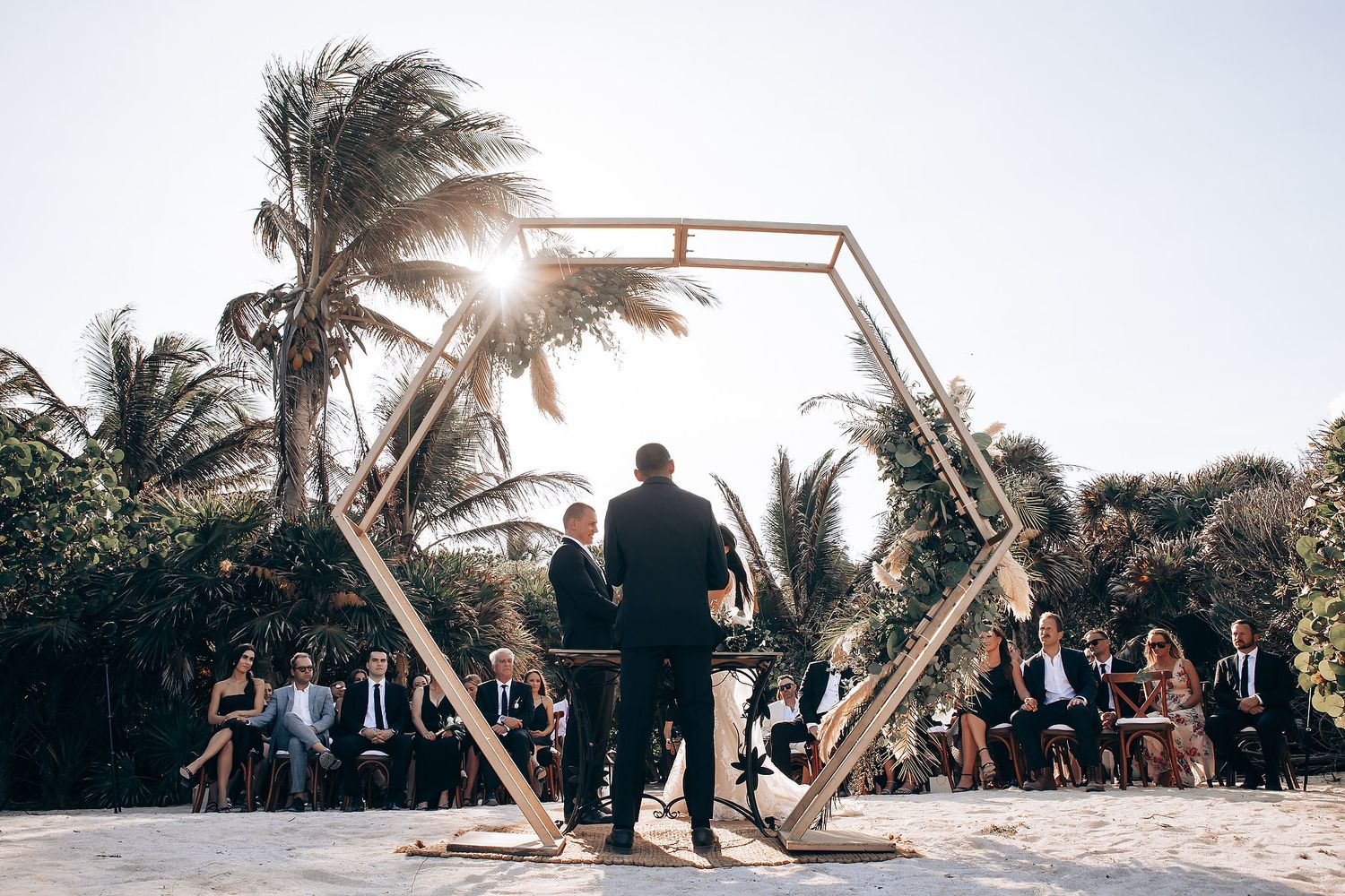 Beachfront wedding ceremony setup with wooden arch and chairs against palm tree backdrop.