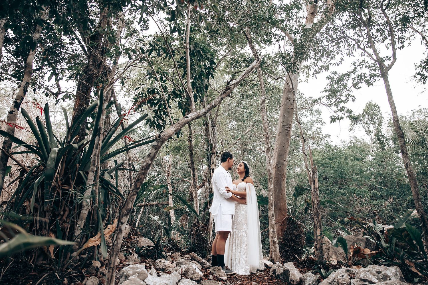A romantic couple in white outfits embraces among tall trees in a serene forest setting during a photo session.
