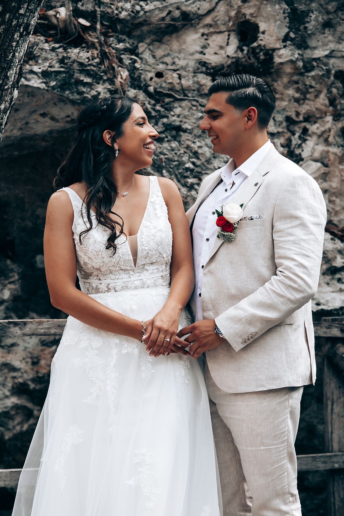 A romantic wedding couple in white attire share intimate moments against a stone wall backdrop.