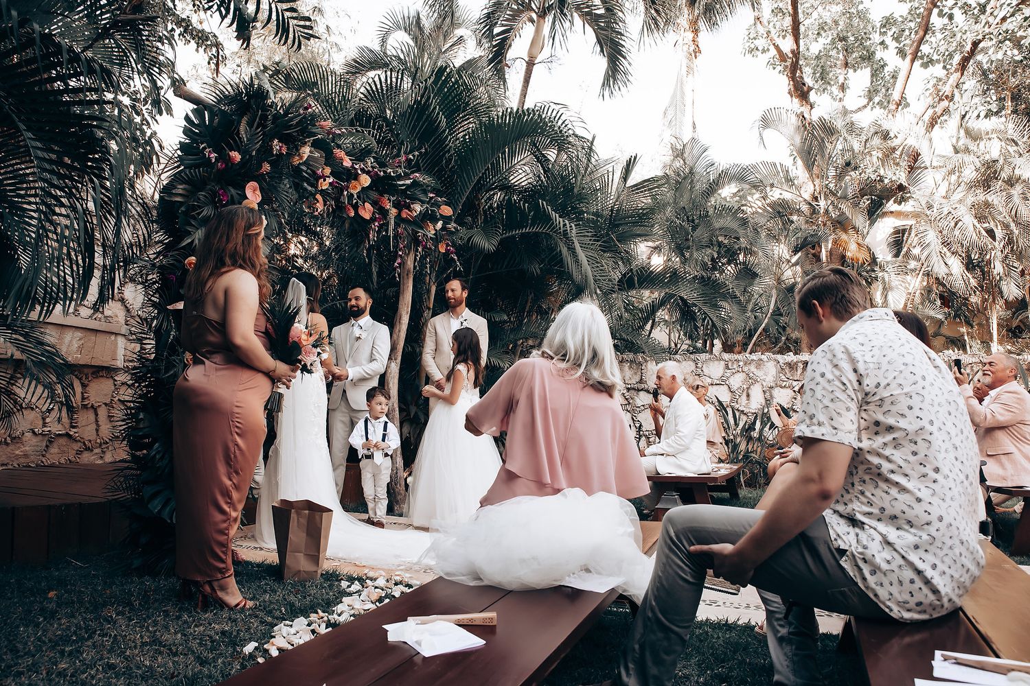 Wedding guests gather outdoors under palm trees for a casual celebration during a daytime ceremony.