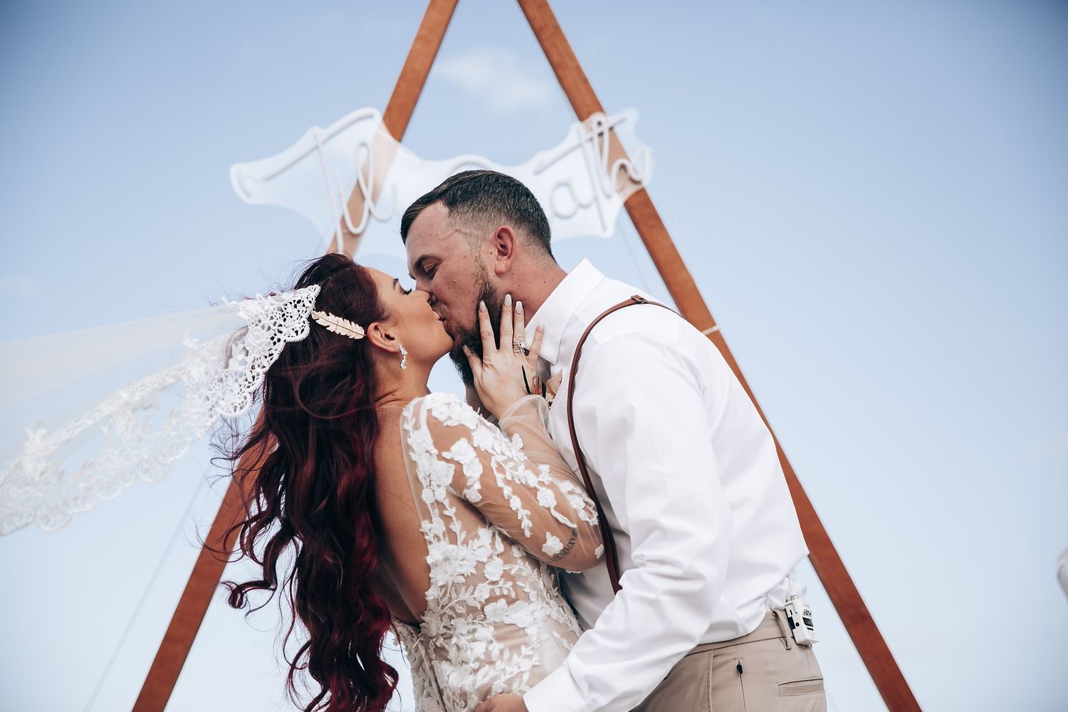 A romantic wedding ceremony under a triangular wooden arch on a tropical beach at sunset.