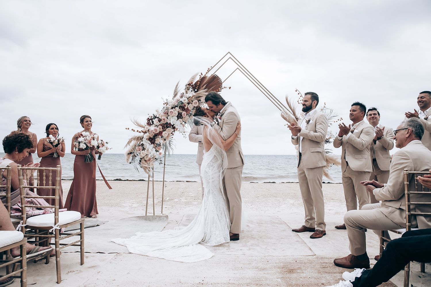 Wedding ceremony setup on beach with white chairs and floral arrangements under bright sky.