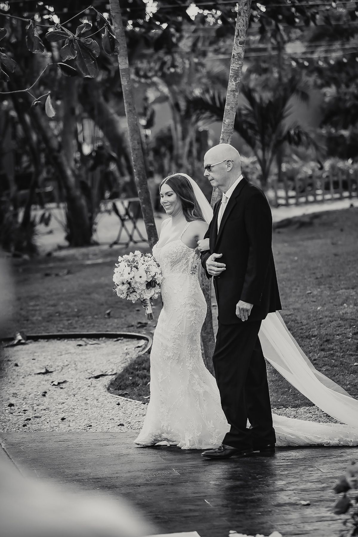 Bride in lace gown with long veil walks down aisle with escort at elegant outdoor wedding ceremony.