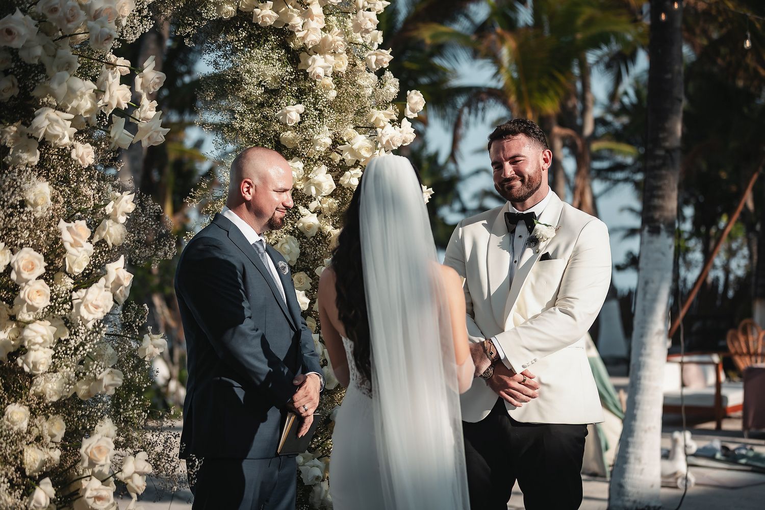 Bride and groom exchanging vows at an outdoor tropical wedding ceremony beneath a floral arch of white roses.