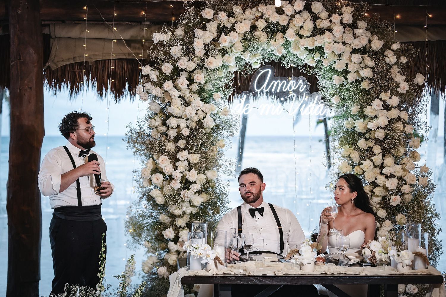 A groom and bride sit at a sweetheart table beneath a floral arch with an Amor sign as a guest gives a speech.