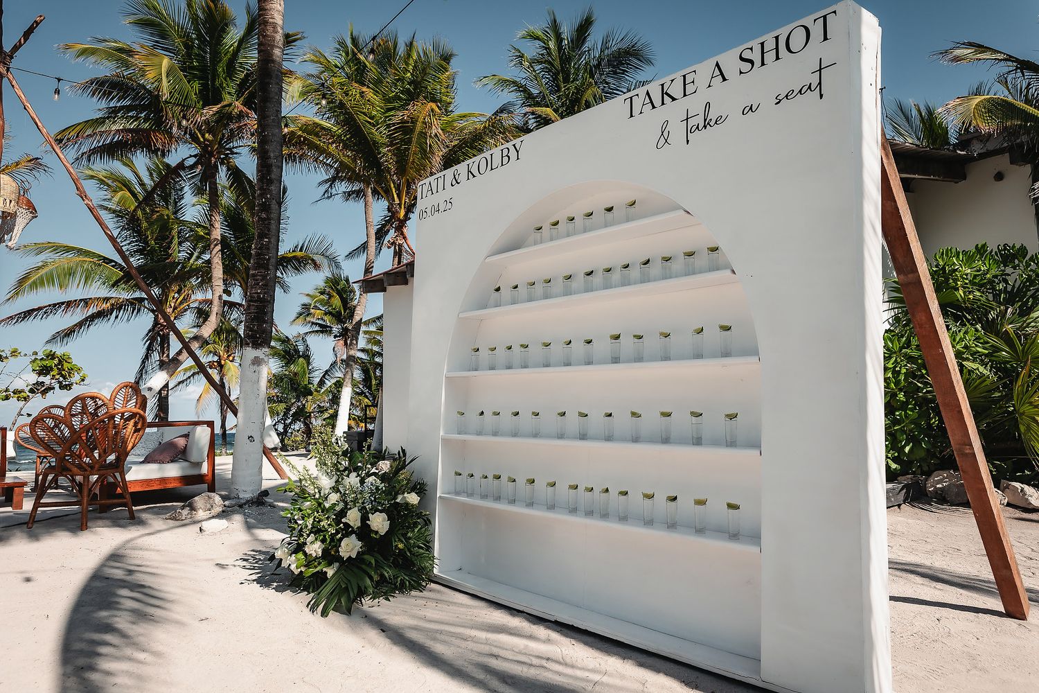White arched shot wall display on beach with 'Take a Shot' sign, surrounded by palm trees and floral arrangements.
