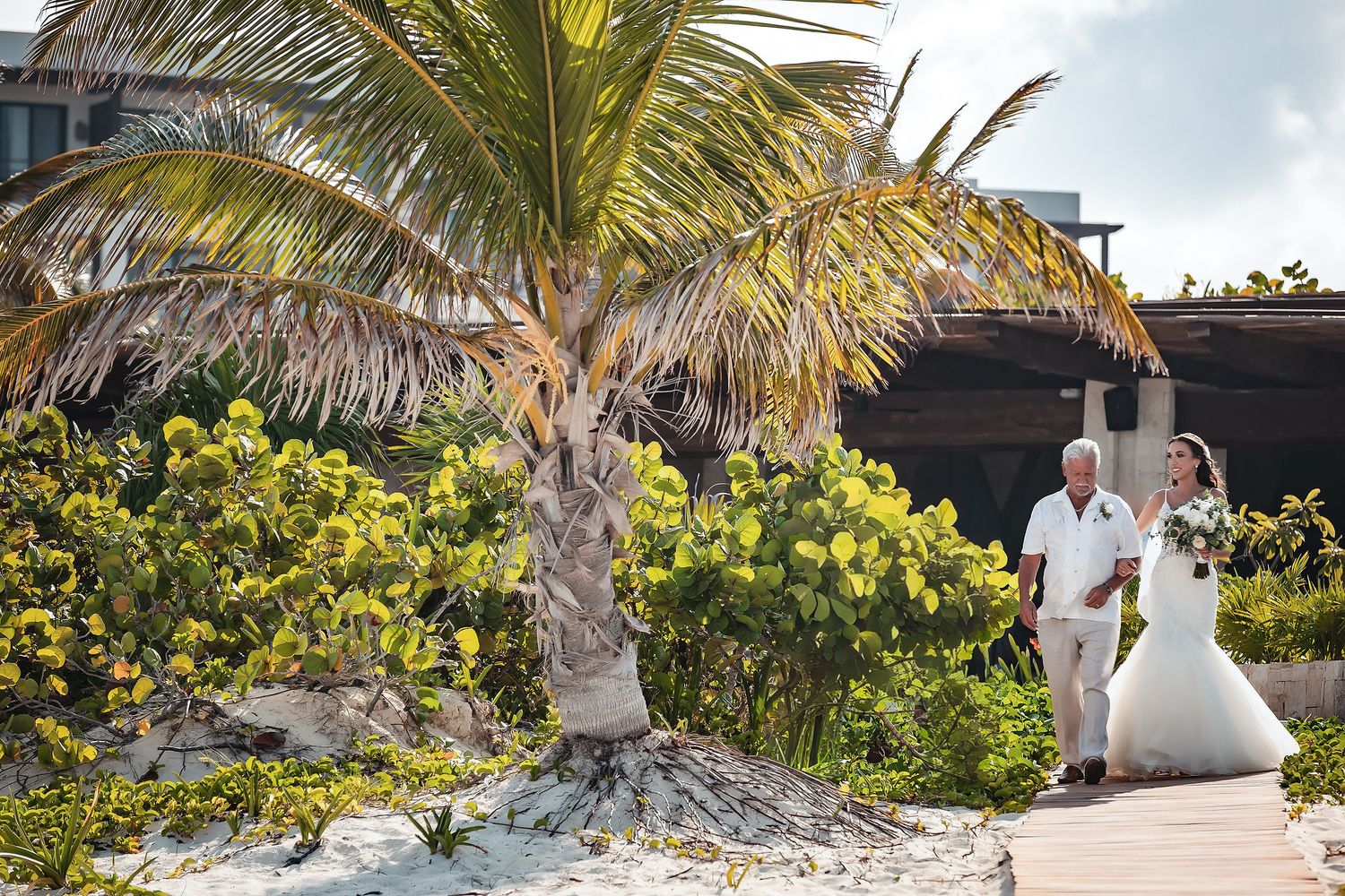 Bride and groom walking on a sandy path surrounded by tropical palms at a beachside wedding venue.