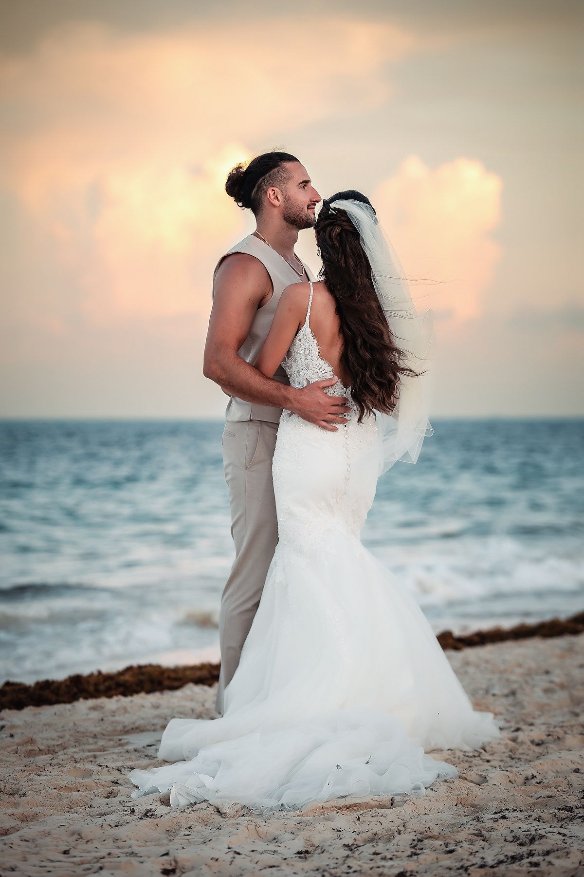 Bride and groom share a romantic kiss on a sandy beach at sunset, with ocean waves in the background.