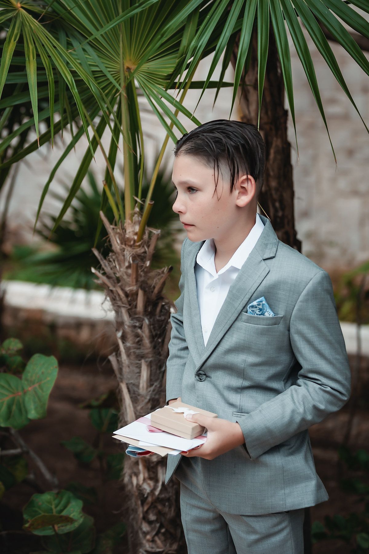 Boy in gray suit holding book stands near tropical palm tree outdoors.
