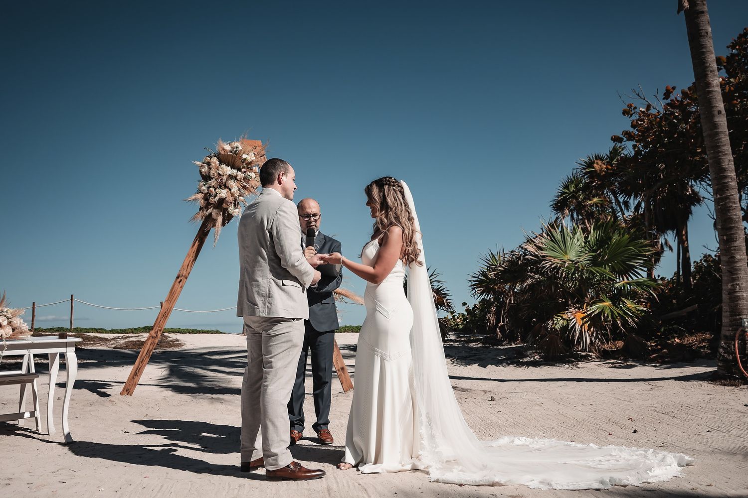 Couple exchanging vows on a sunny beach with a floral arch and officiant.