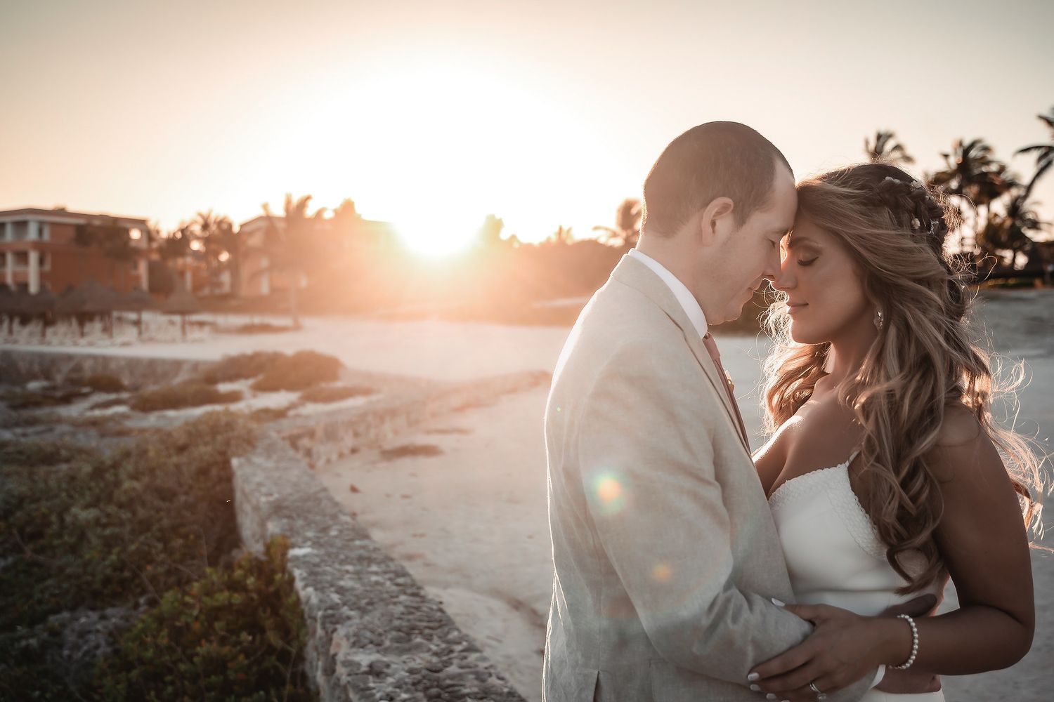 Bride and groom share a romantic sunset embrace on a tropical beach, golden light glowing behind them.