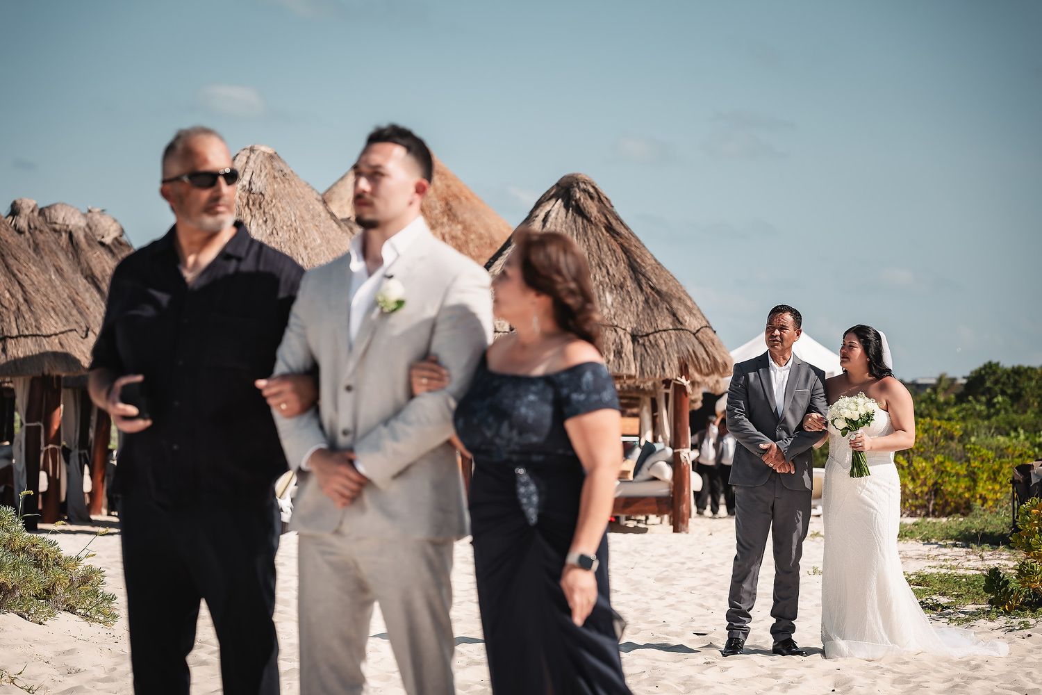 Bride and groom with family on sandy beach at tropical destination wedding ceremony with thatched huts.