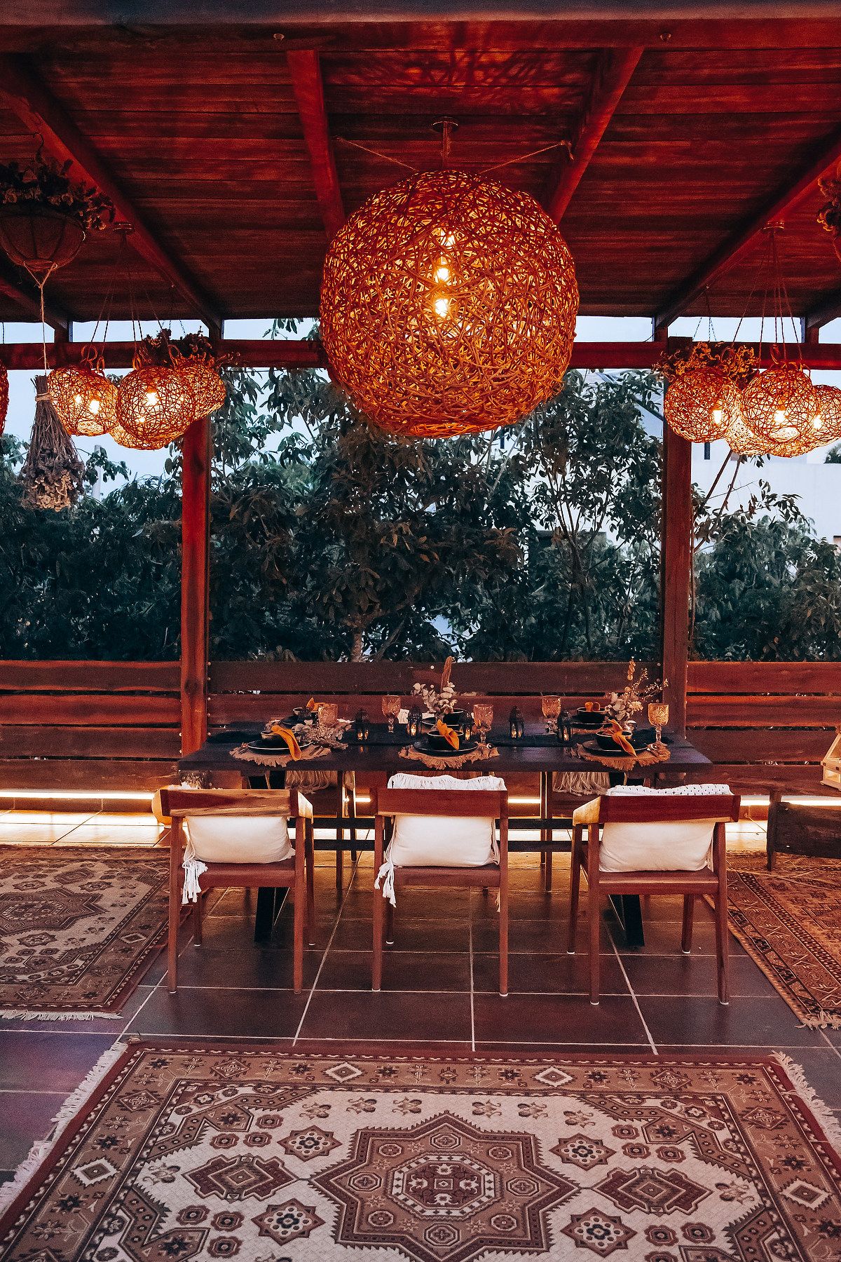 Warm bohemian outdoor dining setup with woven rattan globe pendant lights over rustic wooden table.