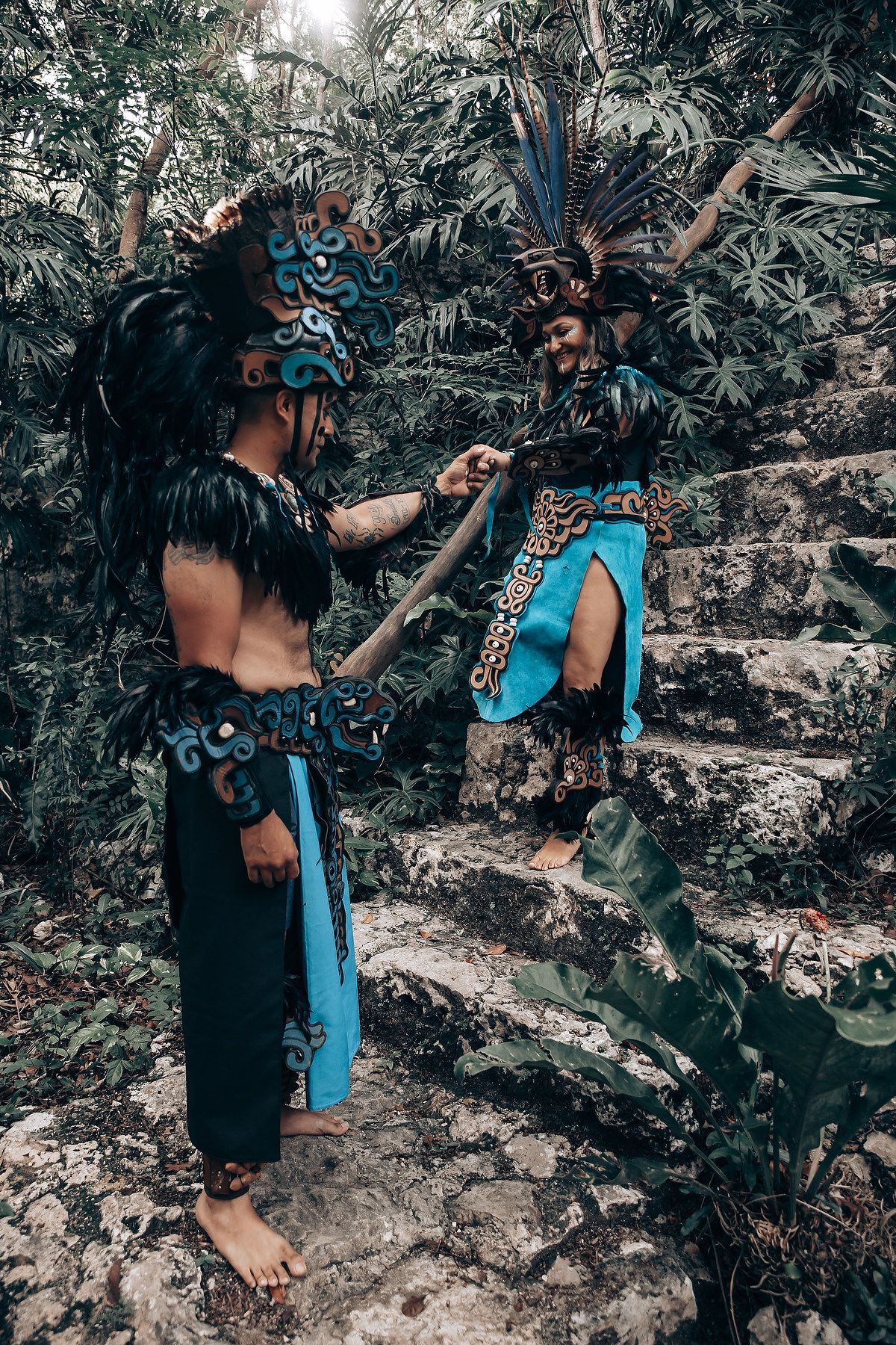 Two performers in elaborate Mayan costumes with feathered headdresses on ancient stone steps in a jungle.