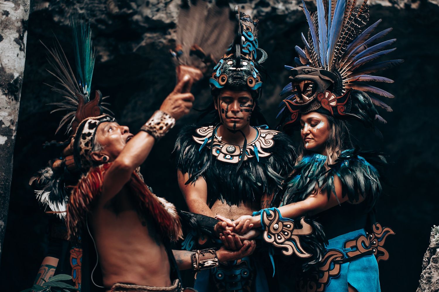 Three performers in elaborate Aztec ceremonial costumes with feathered headdresses and face paint performing a traditional ritual.