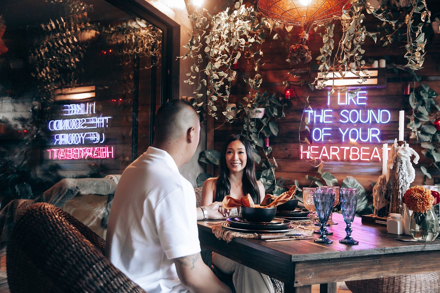 Couple dining at a romantic bohemian restaurant with neon sign reading I Like The Sound Of Your Heartbeat on wooden wall.