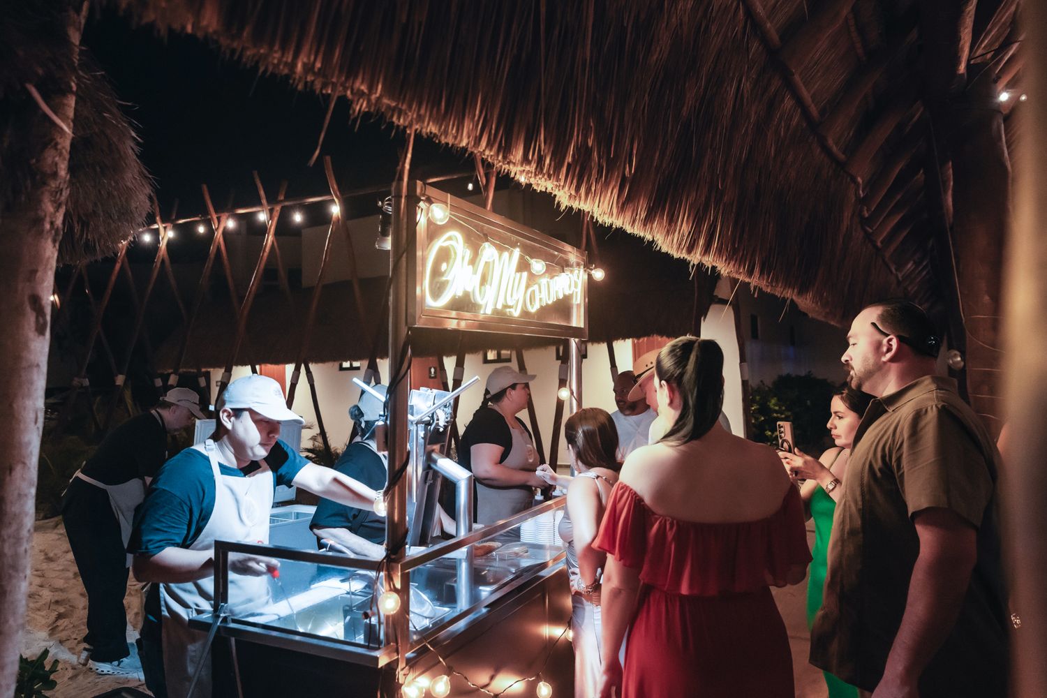 Guests ordering at a thatched-roof outdoor bar with neon sign at a tropical night event.