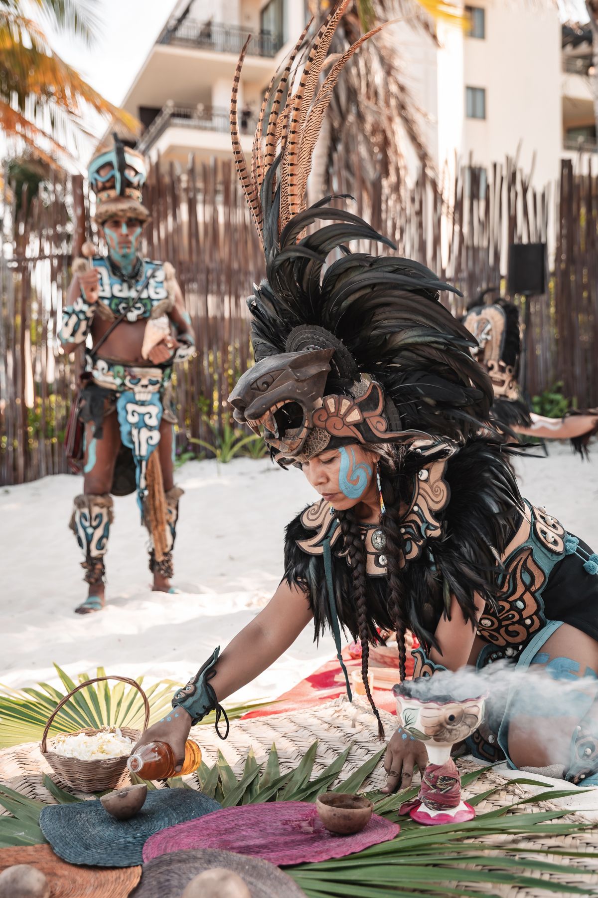 Mayan performers in traditional feathered headdresses and body paint conduct a ceremonial ritual on a sandy beach.