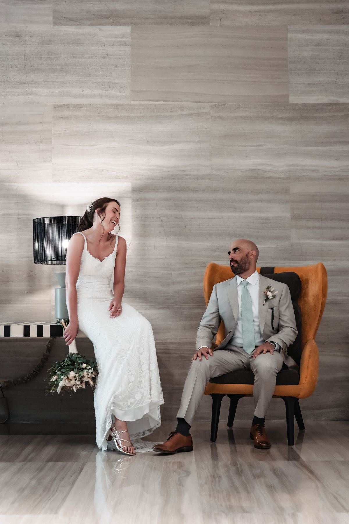 Bride in white gown and groom in gray suit sitting together laughing in an elegant modern venue.