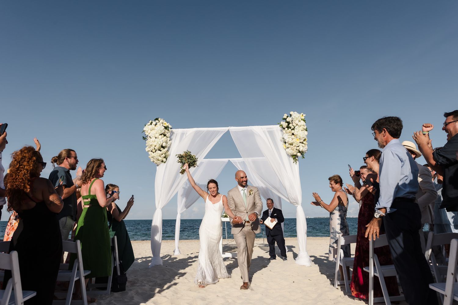 Joyful newlyweds walk down beach aisle under white floral arch, guests clapping, clear blue sky backdrop.