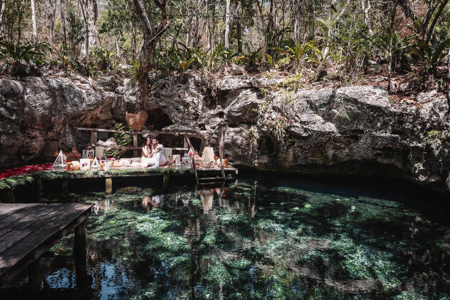 A couple enjoys a romantic picnic setup on a wooden dock beside a crystal-clear turquoise cenote surrounded by jungle rocks.