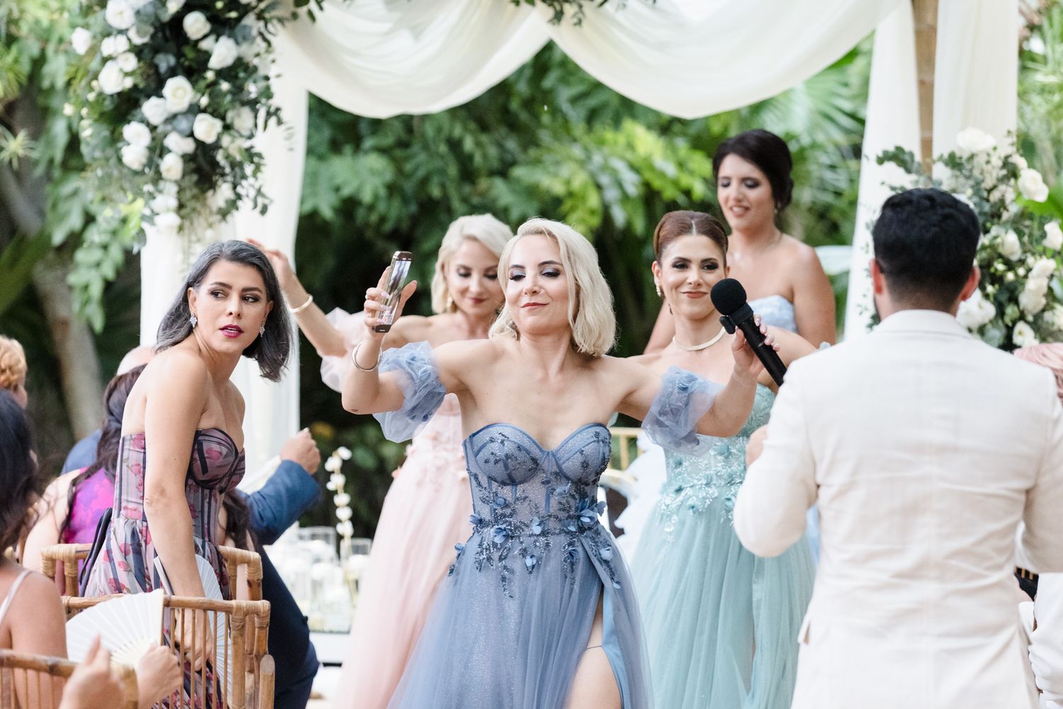 Bridesmaids in blue floral gowns celebrate at an outdoor wedding ceremony surrounded by lush greenery and white floral decor.