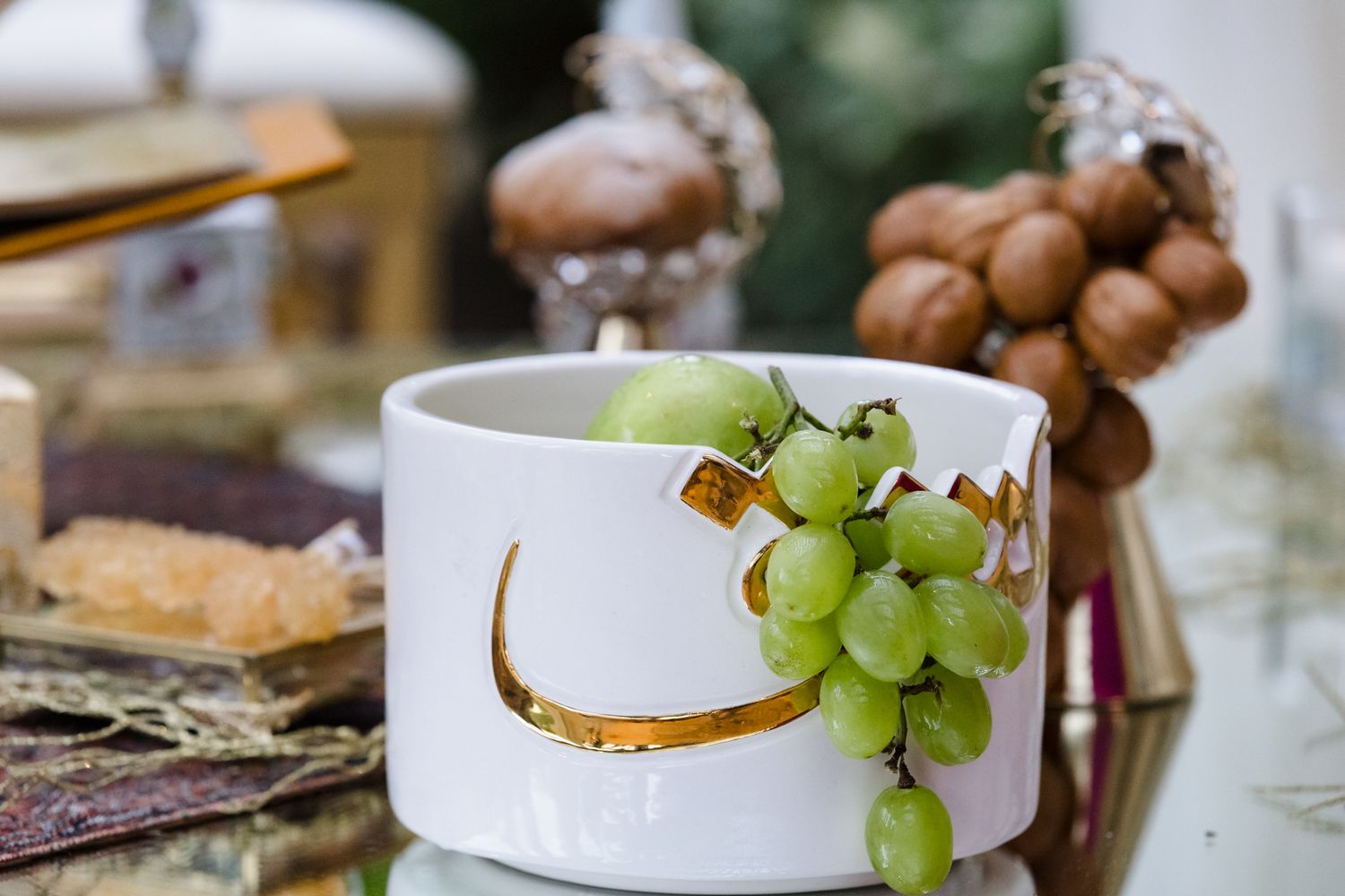White ceramic bowl with gold Arabic calligraphy holding fresh green grapes, surrounded by nuts and food items on a table.