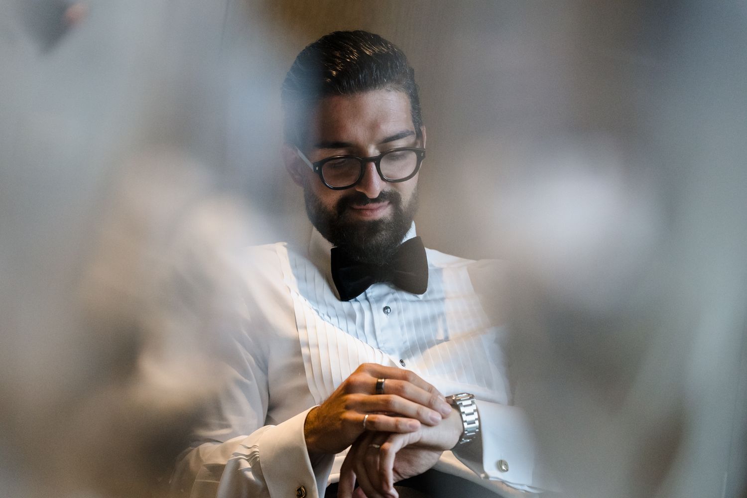 A well-dressed man in a white tuxedo and black bow tie adjusts his cufflinks, photographed through a soft bokeh foreground.