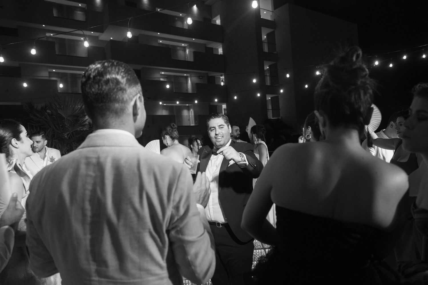 Black and white photo of wedding guests dancing and socializing at an outdoor evening reception with string lights.