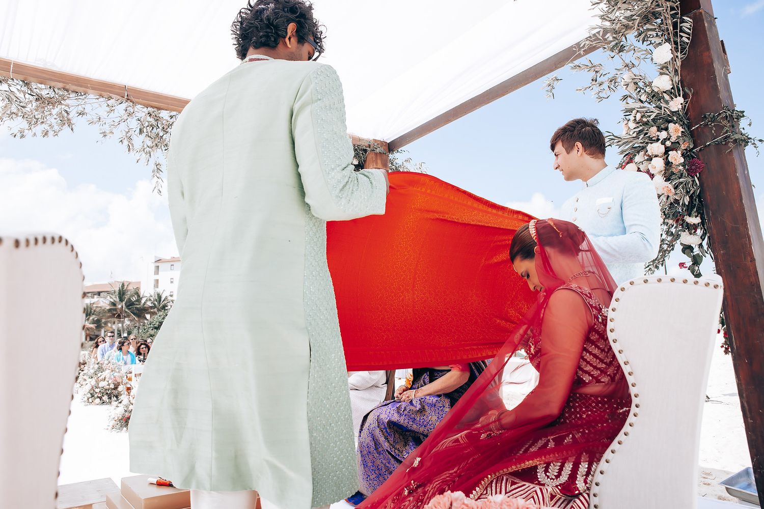 Wedding ceremony participants in traditional red and white attire against a beachfront backdrop.