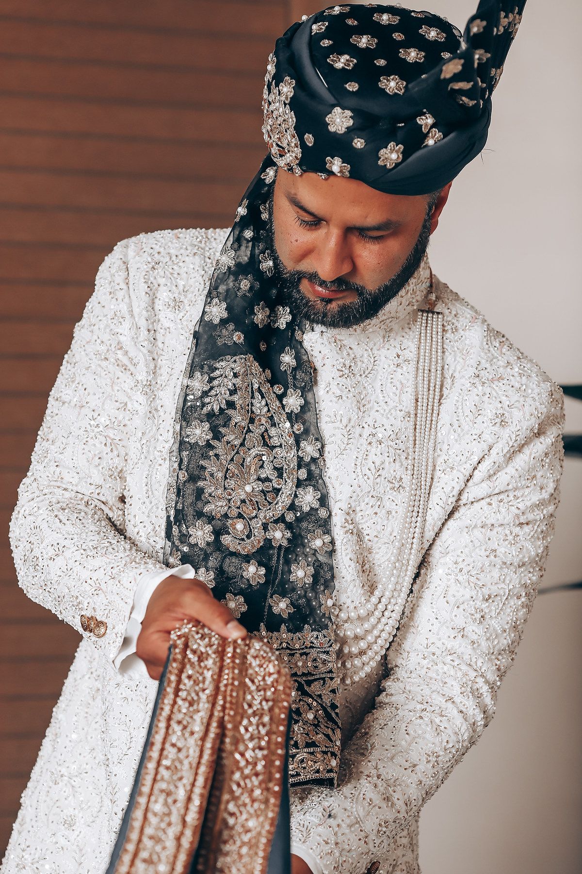 A person in an embellished white sherwani and floral headpiece adjusts traditional wedding accessories.