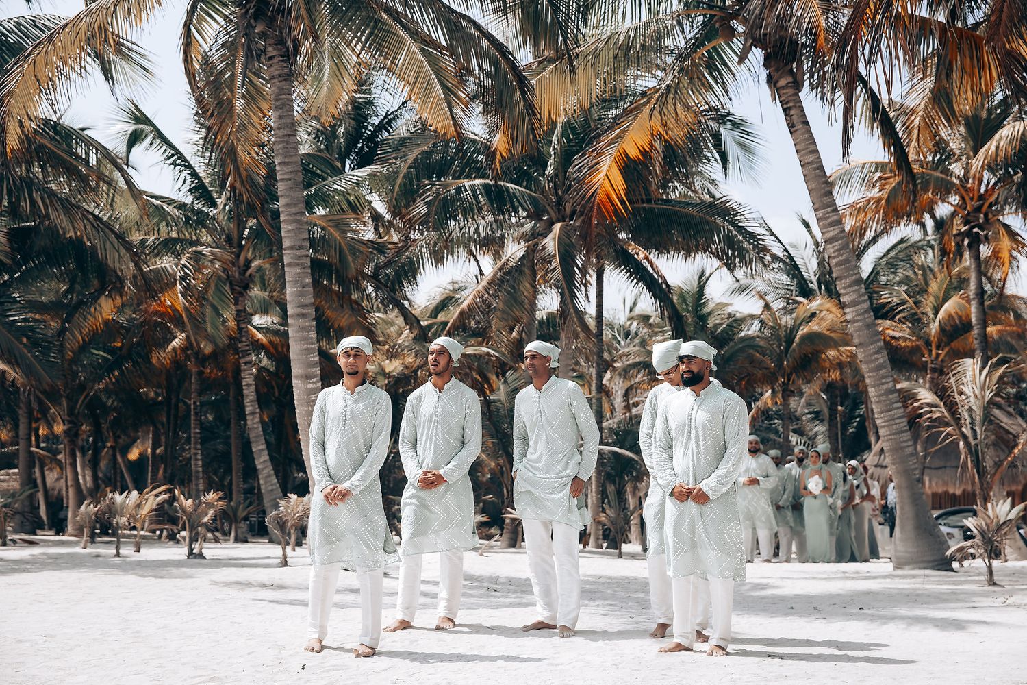 Wedding party in all-white attire poses along tropical beach with palm trees.