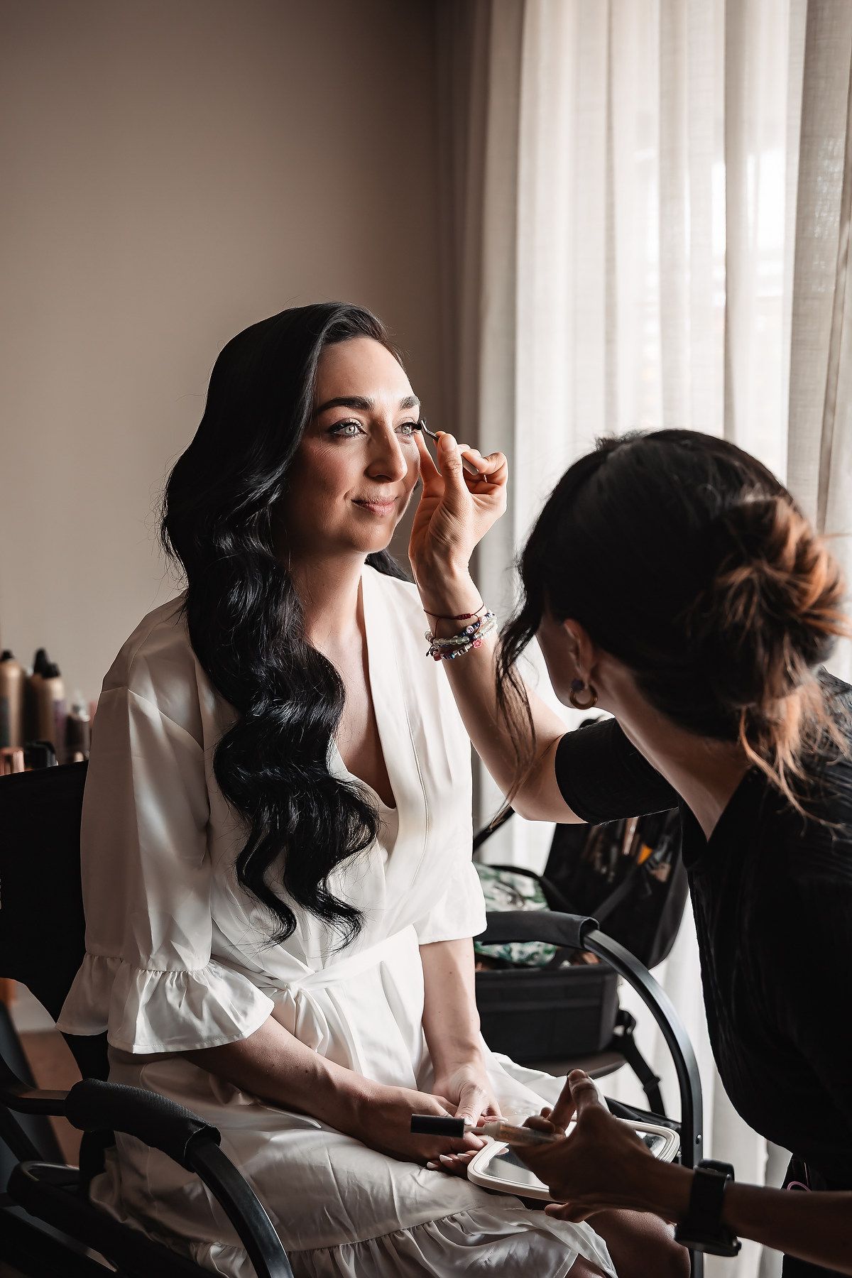 Makeup artist applies cosmetics to woman in white robe sitting in chair by window during bridal preparation.
